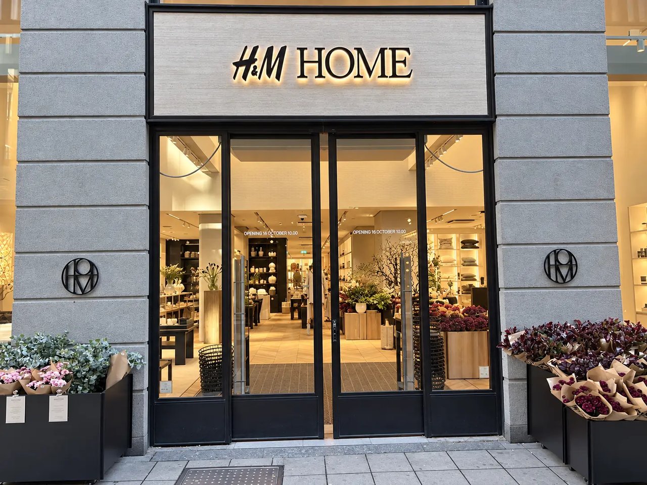 H&M HOME:s butiksfasad på Drottninggatan 50 i Stockholm med upplyst skylt och blomsterarrangemang vid entrén.
